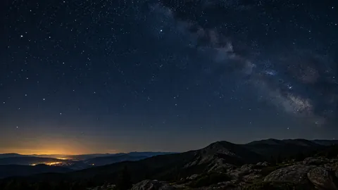 Editorial illustration of a starry sky above dark mountain ridges with distant town lights near the horizon.